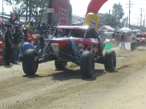 1990 Ford Bronco - red tube-frame off-road buggy goes airborne over a dirt jump as spectators watch under a red Tecate inflatable arch.