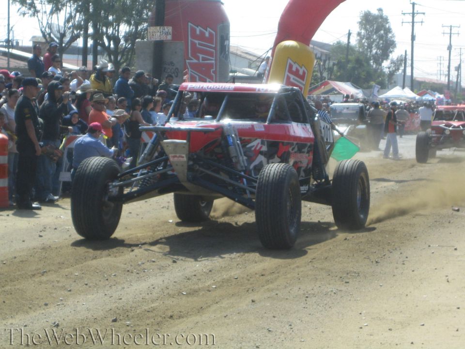 Red tube-frame off-road buggy airborne on a dirt track with crowd behind