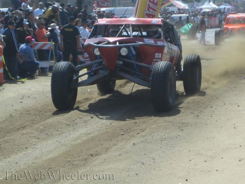 1990 Ford Bronco - dark red tube-frame off-road buggy kicks up dust as it lands on the dirt course in front of a large spectator crowd.