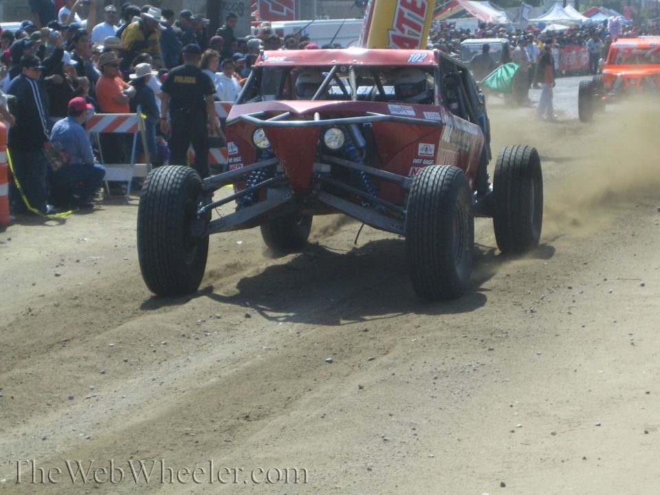 Dark red tube-frame off-road buggy landing on a dirt course before a crowd