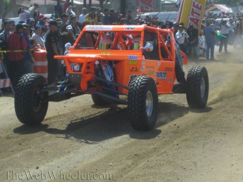1990 Ford Bronco - bright orange tube-frame off-road race buggy covered in sponsor decals races along a dusty dirt track past onlookers.
