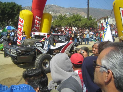 1990 Ford Bronco - Spectators crowd around the No. 106 off-road buggy near the red Tecate start arch at a hillside desert race.