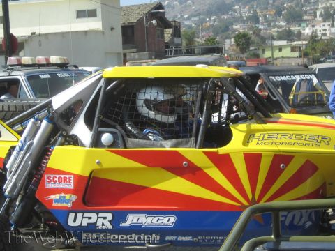 1990 Ford Bronco - helmeted driver sits in the caged cockpit of a yellow and red Herder Motorsports off-road buggy with SCORE decals.