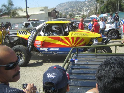 1990 Ford Bronco - yellow and red No. 110 off-road buggy with sunburst graphics sits among spectators at a staging area.