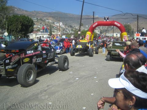 1990 Ford Bronco - Off-road race vehicles including No. 111 stage on a town street below a red inflatable start arch as crowds line the route.