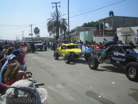 1990 Ford Bronco - row of off-road buggies including the black No. 113 stages on a street lined with palm trees and spectators.