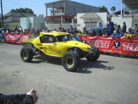 1990 Ford Bronco - bright yellow CATTRAC off-road buggy rolls down a street lined with red Tecate banners and spectators.