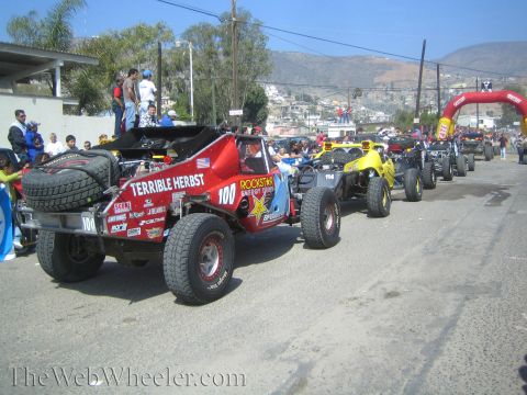 1990 Ford Bronco - line of off-road buggies led by the red No. 100 Terrible Herbst car queues on a street with a hillside town behind.