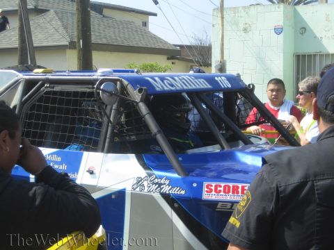1990 Ford Bronco - blue McMillin No. 116 off-road buggy with a caged cockpit and SCORE International decal among officials and onlookers.