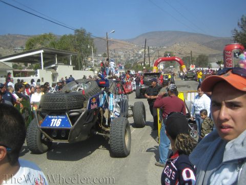 1990 Ford Bronco - No. 116 off-road buggy sits on a street as a young spectator looks on, with a red start arch and hillside town behind.