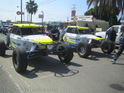 1990 Ford Bronco - Two yellow and white Wilson-sponsored off-road buggies sit side by side on a street with crew and shops behind.