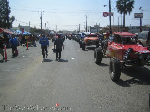 1990 Ford Bronco - Off-road buggies stage along a wide street lined with palm trees and vendor tents as people walk the race route.