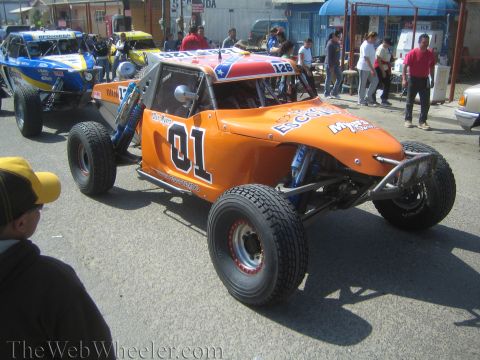 1990 Ford Bronco - orange No. 01 off-road buggy with a Texas-flag roof panel sits on a street among other buggies and onlookers.