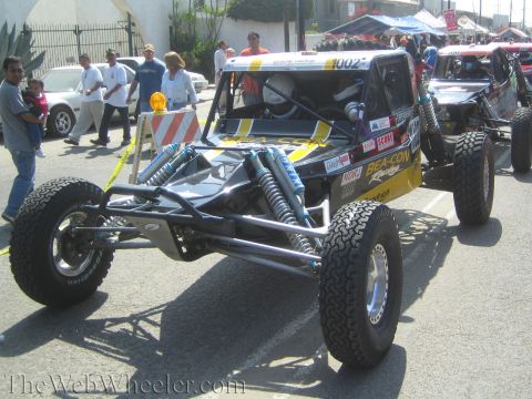 1990 Ford Bronco - black and silver long-travel tube-frame off-road buggy sits on pavement at a staging area with bystanders nearby.
