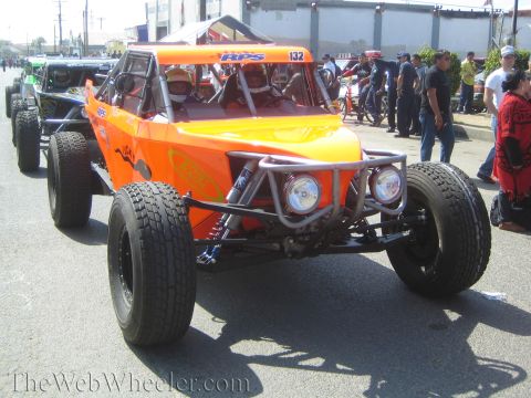 1990 Ford Bronco - orange No. 132 off-road buggy with round headlights and a driver in the cockpit rolls along a paved staging street.