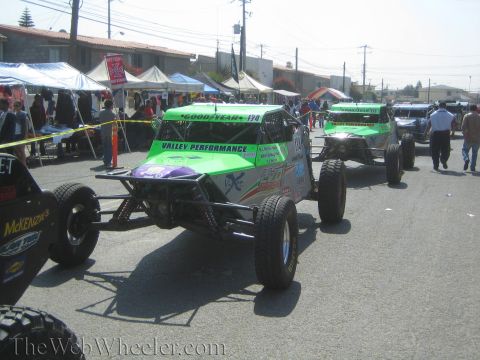 1990 Ford Bronco - Green and silver No. 170 Valley Performance off-road buggies move along a street past vendor tents and spectators.