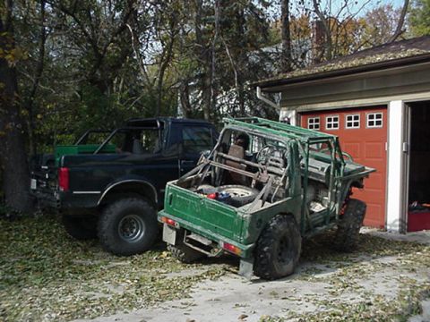 1992 Ford Bronco - Two vehicles, a green off-road vehicle and a black pickup truck, are parked outside a garage on a fall day.