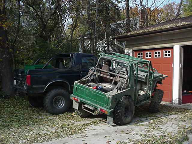 Green off-road vehicle and black pickup truck parked on leaf-covered ground near a garage