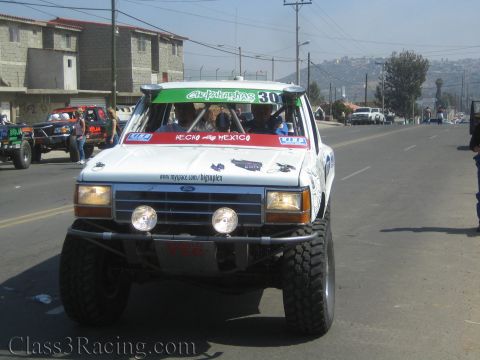 1990 Ford Bronco - Front view of a white Ford off-road race truck numbered 301 with auxiliary lights and a roof sun-strip on a Baja street.