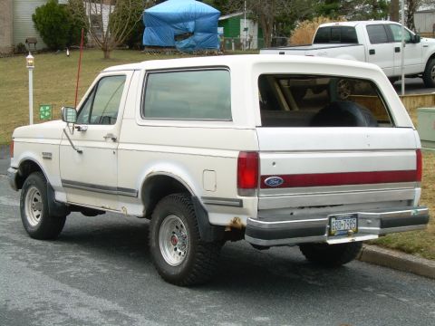 1988 Ford Bronco - The rear of a white Ford Bronco SUV is shown parked on a street with a white pickup truck in the background.