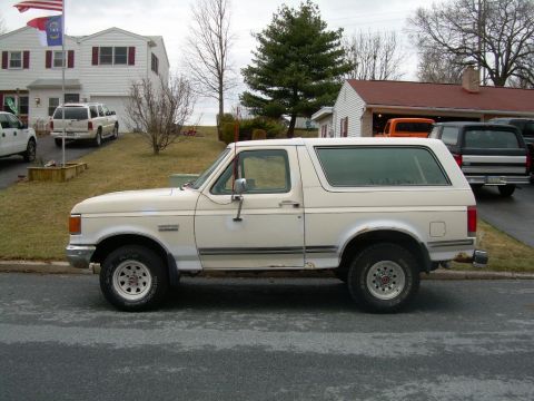 1988 Ford Bronco - A white Ford Bronco is parked on a street, with houses and other vehicles in the background.