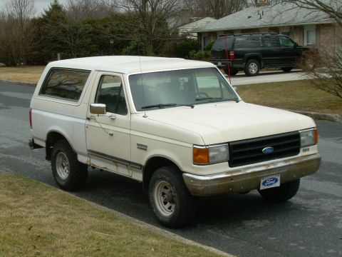 1988 Ford Bronco - A white Ford Bronco is parked on the side of the road, with a black SUV visible in the driveway behind it.