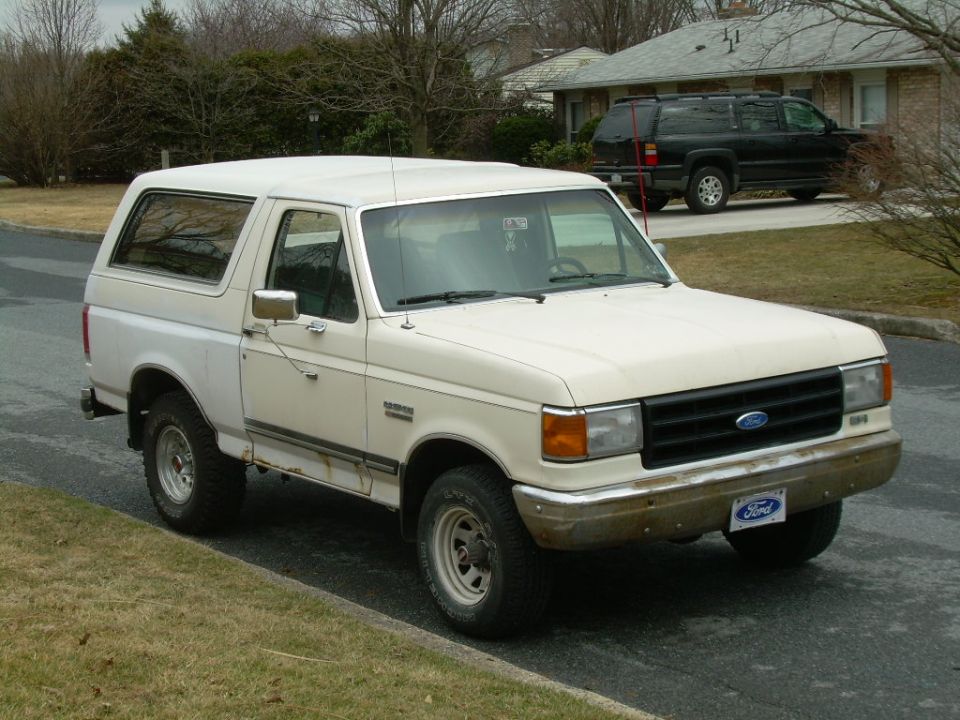 White Ford Bronco parked on a street, showing rust on the lower body panels and a black SUV in the background