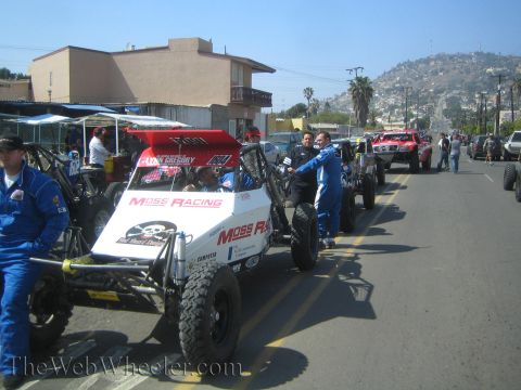 1990 Ford Bronco - Open-wheel off-road race buggies, including a Moss Racing car, staged in a line along a sunny street with crew in suits.