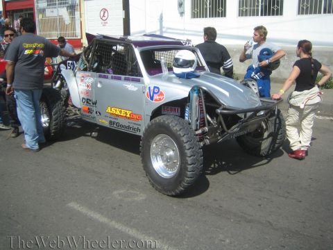 1990 Ford Bronco - silver open-wheel off-road race buggy with a helmeted driver and BFGoodrich livery, surrounded by people on a street.