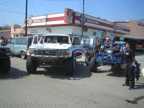1990 Ford Bronco - white Ford prerunner race truck and a blue off-road buggy parked in front of a Gallo Ferreteria hardware store.