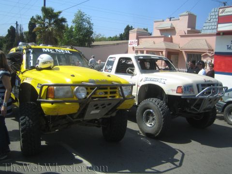 1990 Ford Bronco - yellow Ford off-road race truck numbered 711 sits beside a white prerunner truck on a sunny street.