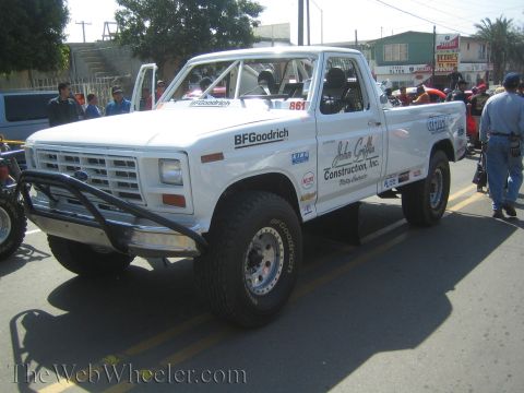 1990 Ford Bronco - white Ford pickup off-road race truck numbered 861 with a caged bed and BFGoodrich livery on a paved street.
