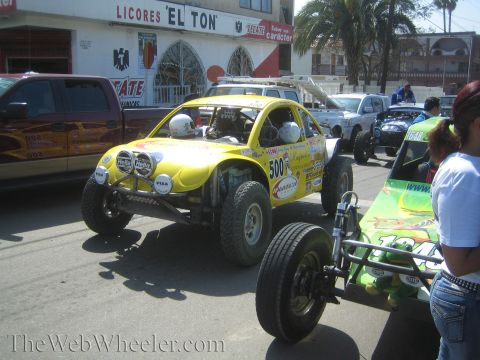 1990 Ford Bronco - yellow open-wheel off-road race buggy numbered 500 parked in front of a Licores El Ton liquor store with spectators.