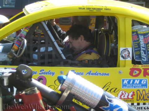 1990 Ford Bronco - Close view into a yellow off-road race car cockpit showing roll cage, harness and a seated driver, with a coil-over shock in front.