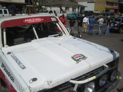 1990 Ford Bronco - High angle over the hood and red roof banner of off-road race truck 303, with crew members gathered in the street behind.