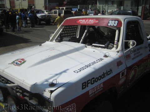 1990 Ford Bronco - Side-front view of white off-road race truck 303 showing the long hood with BFGoodrich and Bilstein decals on a busy street.