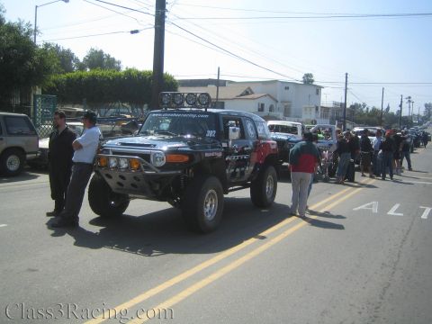 1990 Ford Bronco - black Toyota FJ Cruiser off-road race truck numbered 302 with roof light bar and lift, staged on a street with spectators.