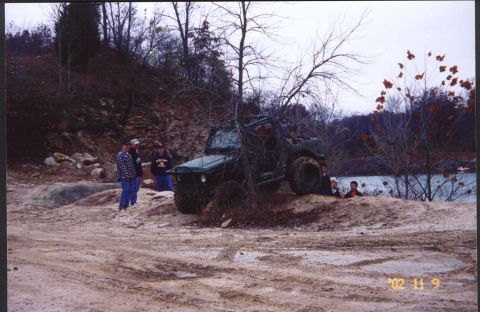 1992 Ford Bronco - An off-road vehicle navigates a muddy trail near a body of water, surrounded by trees and rocky terrain.