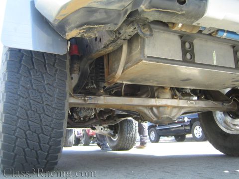 1990 Ford Bronco - Low-angle undercarriage shot of a lifted off-road truck revealing the driveshaft, blue shock absorbers and chunky all-terrain tire.