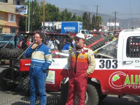 1990 Ford Bronco - Two drivers in colorful racing suits stand before a white and red race truck numbered 303 amid a crowd of spectators.