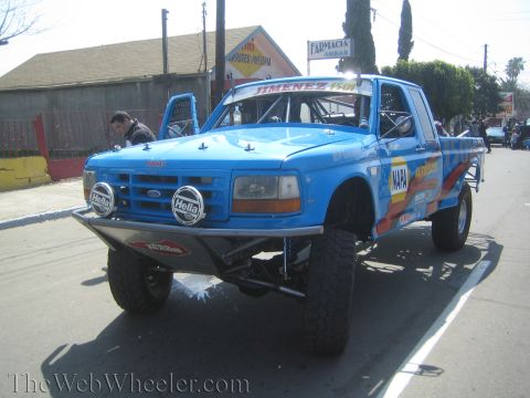 1990 Ford Bronco - Front three-quarter view of a lifted blue Ford race truck with Hella lights, tube bumper and Jimenez and NAPA decals.