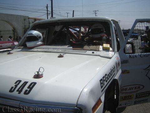 1990 Ford Bronco - white off-road race truck numbered 349 with a helmet resting on the hood, exposed roll cage and BFGoodrich and Deaver decals.
