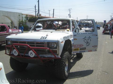 1990 Ford Bronco - white lifted off-road race truck numbered 349 with red tube bumper and open doors moving down a sunny street.