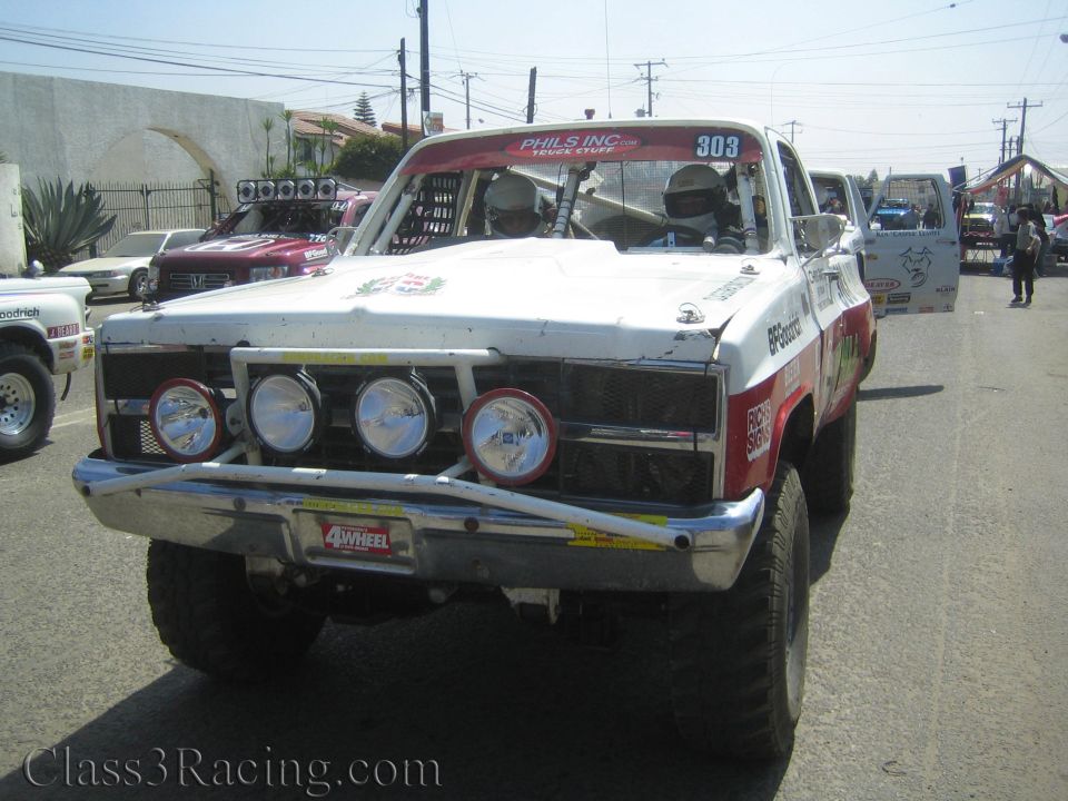 White and red off-road race truck number 303 with four front light pods