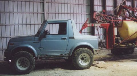 1992 Ford Bronco - A blue Suzuki Samurai is parked indoors next to a large piece of farm machinery.