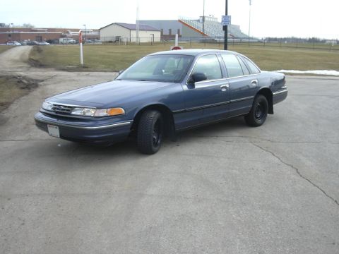 1996 Ford Crown Victoria - A blue sedan is parked on a paved surface with a building and stadium in the background.