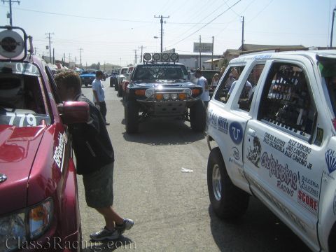 1990 Ford Bronco - Off-road race trucks queued on a street, a black Toyota FJ Cruiser in the middle flanked by a maroon truck and a white SUV.