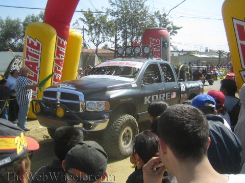 1990 Ford Bronco - black Dodge Ram off-road race truck with roof lights passes under an inflatable Tecate arch as a crowd watches.