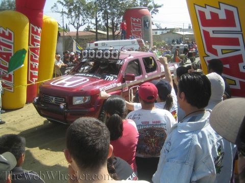 1990 Ford Bronco - red Honda Ridgeline race truck numbered 779 rolls under an inflatable Tecate arch surrounded by cheering spectators.