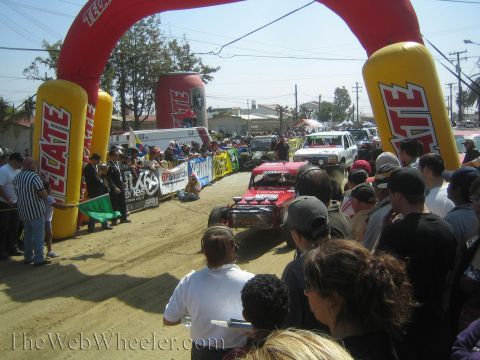 1990 Ford Bronco - race truck kicks up dust launching from a dirt start line beneath an inflatable Tecate arch as crowds line the course.